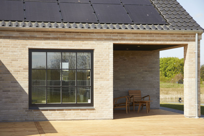 Facade fra eurodan-huse med lyst murværk, sort sprosset vindue, overdækket terrasse, træmøbler og tagflade med integrerede solceller – inspiration til nybyggeri, husbyggeri og husdrømme med klassiske vinduesdetaljer.