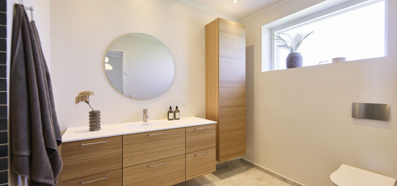 Badeværelse fra eurodan-huse med langt træmøbel, rund spejlflade over vasken, højt skab til opbevaring, væghængt toilet, højtsiddende vindue og mørke håndklæder – inspiration til gennemtænkt nybyggeri og badeværelse i et nybygget hus.