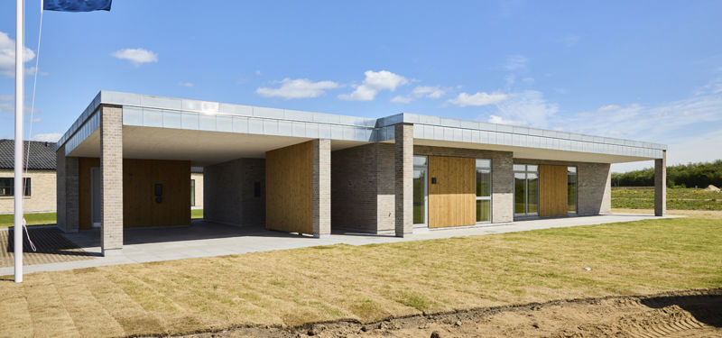 Moderne længehus i nybyggeri med mørk murstensfacade, fladt tag, trædetaljer og integreret overdækning ved indgangen.