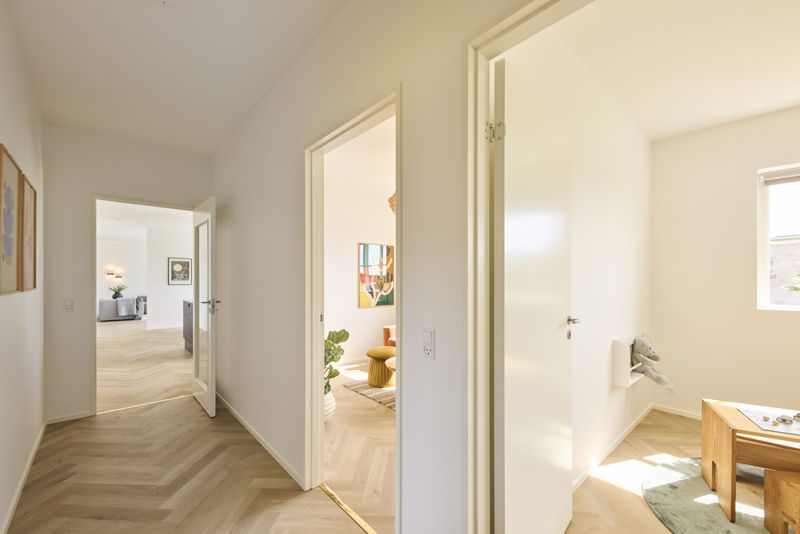Lys gang i udstillingshus fra eurodan-huse med sildebensgulv, åbne døre til stue og børneværelse samt kig til moderne opholdsrum, der viser en gennemtænkt planløsning og inspirerer til nybyggeri, husbyggeri og indretning af et nyt familiehus.