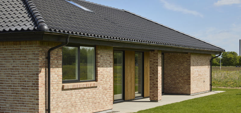 Facade på nybygget udstillingshus fra eurodan-huse med lys murstensfacade, sort tegltag, ovenlysvindue, trædetaljer ved indgang og grøn have som inspiration til husbyggeri