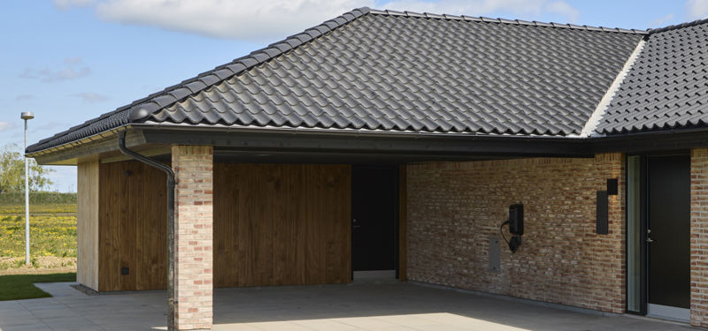 Integreret carport i nybygget udstillingshus fra eurodan-huse med sort tegltag, lys murstensfacade, træbeklædning og overdækket indgang som inspiration til praktisk husbyggeri og moderne husdrømme
