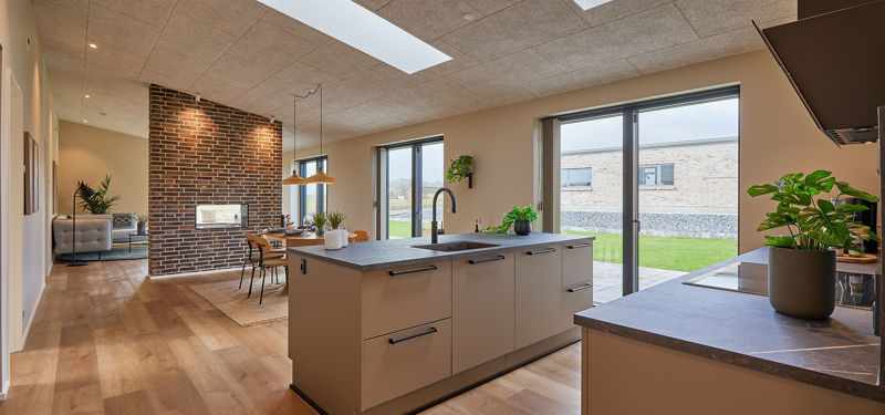Moderne køkken-alrum med stor køkkenø, ovenlysvinduer, lyst trægulv, rå murstensvæg og brede glaspartier mod haven – inspirerende indretning til nybyg og husdrømme.
