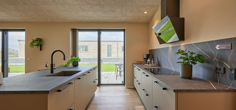 Moderne køkken med lang køkkenø, varme beige fronter, mørke bordplader, sorte greb og armatur, ovenlysvinduer samt glasdør mod haven – stilren inspiration til nybyg og husdrømme.