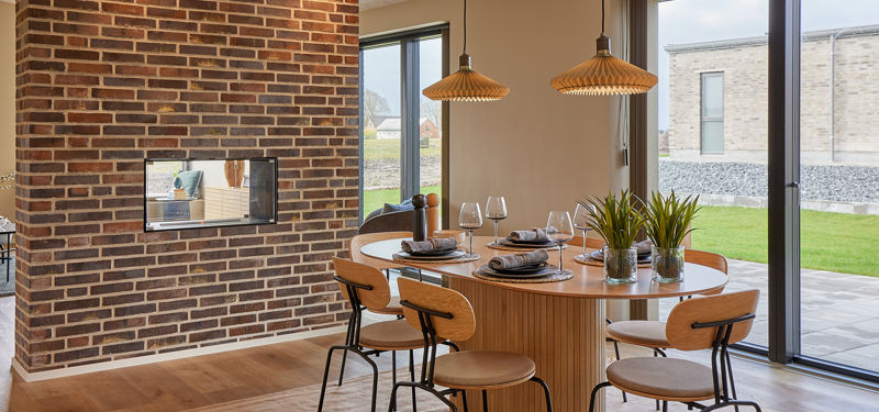 Indbydende spiseplads med rundt træbord, moderne stole, flettede pendellamper, store glaspartier mod haven og rå murstensvæg med indbygget pejs – stemningsfuld inspiration til nybyg og husdrømme.