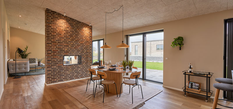 Indbydende spiseplads med rundt træbord, moderne stole, pendellamper, rå murstensvæg med indbygget pejs og store glaspartier mod haven – stemningsfuld inspiration til nybyg og husdrømme.