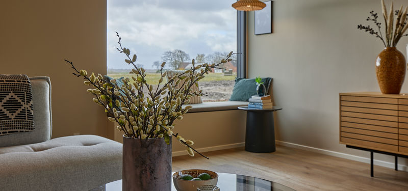 Hyggelig stue med grå sofa, glasbord, stor vase med grene, træskænk og bred vindueskarm indrettet som siddeplads – stemningsfuld inspiration til nybyg og husdrømme.