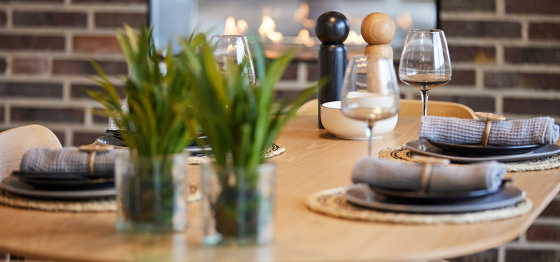 Nærbillede af rundt træbord dækket med tallerkener, stofservietter og vinglas, grønne planter og stemningsfuld pejs i baggrunden – inspiration til nybyg og husdrømme.