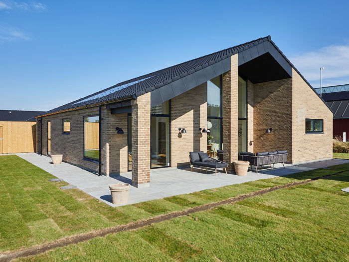 Overdækket terrasse fra eurodan-huse med høj gavl, lyse mursten, sorte tagflader, slanke murstenssøjler, grå terrassefliser og mørke loungemøbler langs facaden – inspiration til arkitektonisk nybyggeri, terrasse og udeliv.