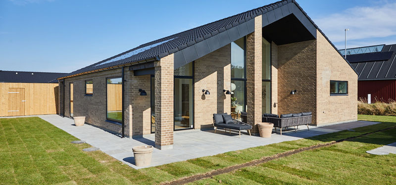 Overdækket terrasse fra eurodan-huse med høj gavl, lyse mursten, sorte tagflader, slanke murstenssøjler, grå terrassefliser og mørke loungemøbler langs facaden – inspiration til arkitektonisk nybyggeri, terrasse og udeliv.