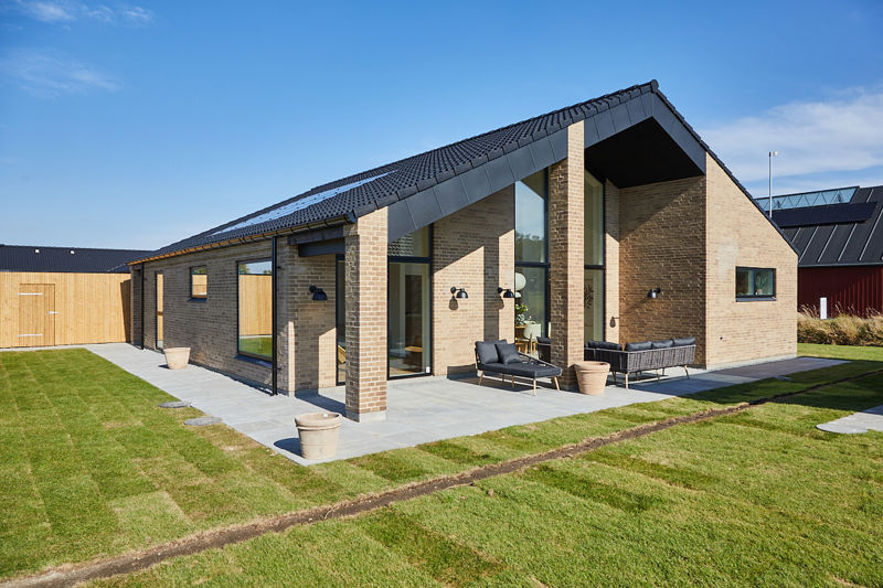 Overdækket terrasse fra eurodan-huse med høj gavl, lyse mursten, sorte tagflader, slanke murstenssøjler, grå terrassefliser og mørke loungemøbler langs facaden – inspiration til arkitektonisk nybyggeri, terrasse og udeliv.