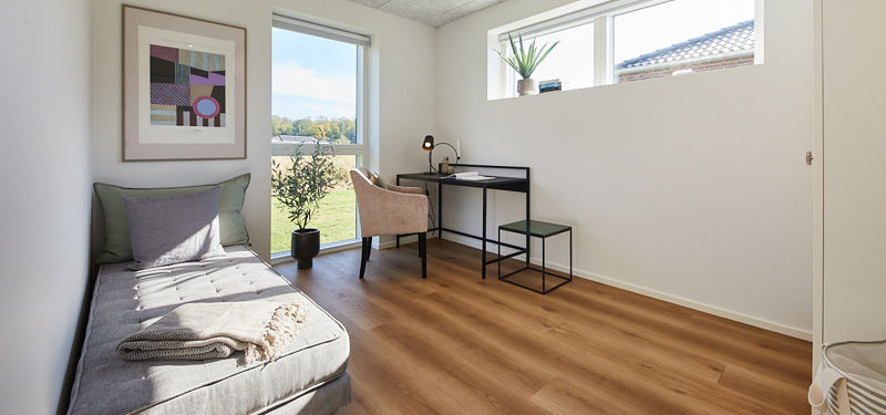 Lyst værelse fra eurodan-huse med grå daybed, skrivebord, rosa stol, grønt skammelsæt, gulv-til-loft-vindue, smalt højtsiddende vindue og plante – inspiration til gæsteværelse, kontor og fleksibelt nybyggeri.