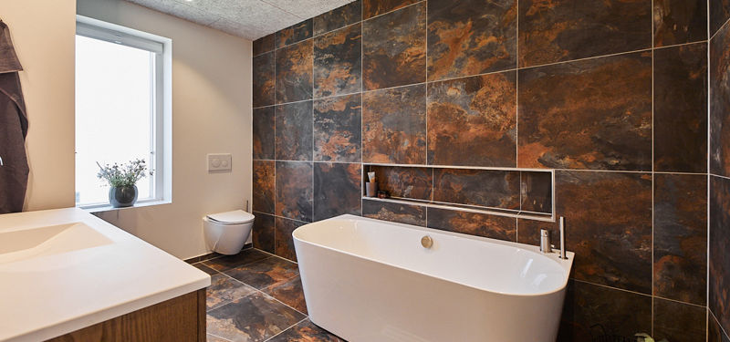 Badeværelse fra eurodan-huse med fritstående hvidt badekar foran mørke rustfarvede fliser, lang indbygget niche, vindue ved toilettet og matchende gulvklinker – konkret inspiration til luksuriøst nybyggeri og moderne husdrømme.
