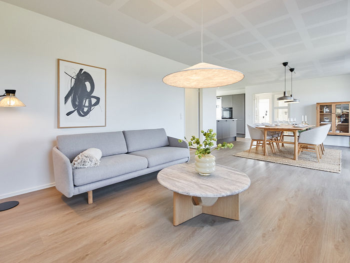 Lyst stueområde fra eurodan-huse med lysegrå sofa, stort rundt marmorlignende sofabord, sort standerlampe, kunstplakat på væggen og direkte kig til spisebord, køkken og vitrineskab – inspiration til nybyggeri, husbyggeri og indretning af åbne opholdsrum.