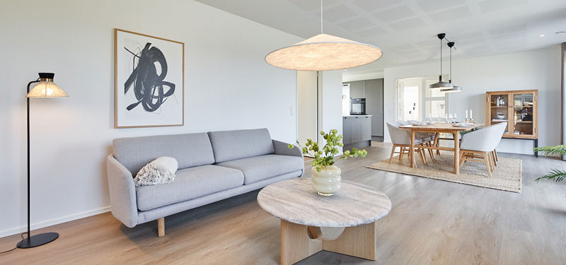 Lyst stueområde fra eurodan-huse med lysegrå sofa, stort rundt marmorlignende sofabord, sort standerlampe, kunstplakat på væggen og direkte kig til spisebord, køkken og vitrineskab – inspiration til nybyggeri, husbyggeri og indretning af åbne opholdsrum.