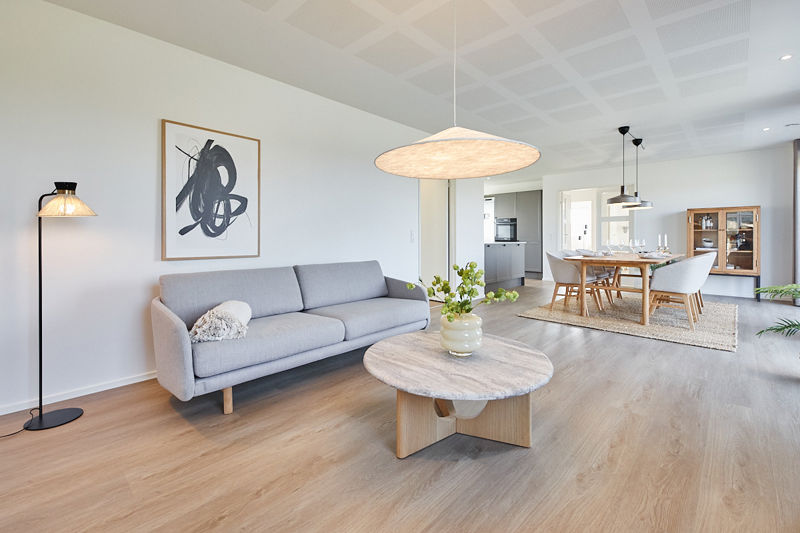 Lyst stueområde fra eurodan-huse med lysegrå sofa, stort rundt marmorlignende sofabord, sort standerlampe, kunstplakat på væggen og direkte kig til spisebord, køkken og vitrineskab – inspiration til nybyggeri, husbyggeri og indretning af åbne opholdsrum.