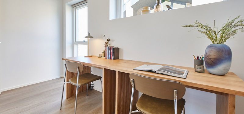 Kontorplads i nybyggeri med langt skrivebord, to arbejdsstationer, høje vinduer og lys indretning til fleksible husdrømme
