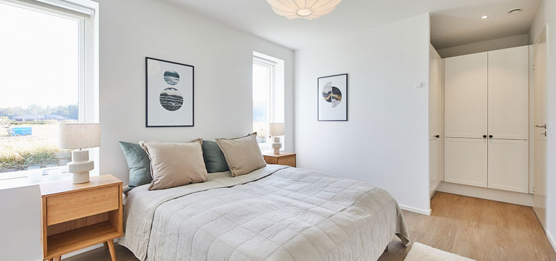 Lyst soveværelse fra eurodan-huse med dobbeltseng, gråt sengetæppe, trænatbord med lampe, to vinduer, enkle billedrammer på væggene og skabsniche med hvide garderobeskabe – inspiration til nybyggeri, husbyggeri og funktionelle husdrømme med naturligt lys.e
