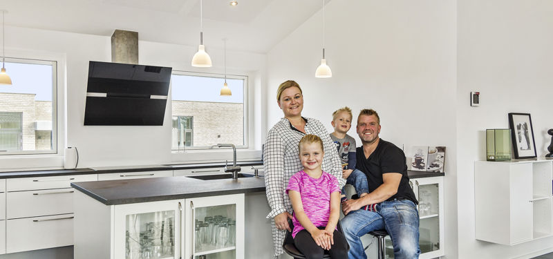 Familie samlet ved køkkenøen i et lyst drømmehus fra eurodan-huse med hvidt inventar, sorte bordplader og loft til kip.
