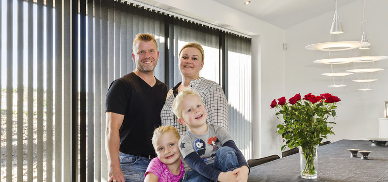 Familie samlet i den lyse spiseafdeling i et drømmehus fra eurodan-huse med store vinduespartier, lamelgardiner og moderne pendler.