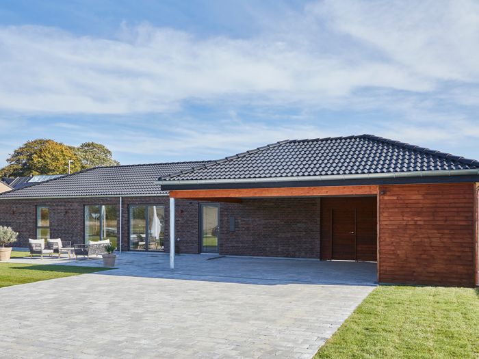 Nybyggeri og husbyggeri med moderne længehus i brunlige mursten, sort tag, træbeklædt carport og overdækket indkørsel fra eurodan-huse