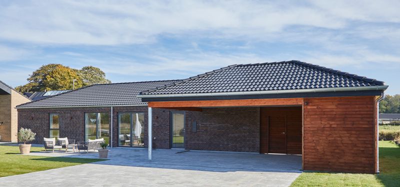 Nybyggeri og husbyggeri med moderne længehus i brunlige mursten, sort tag, træbeklædt carport og overdækket indkørsel fra eurodan-huse