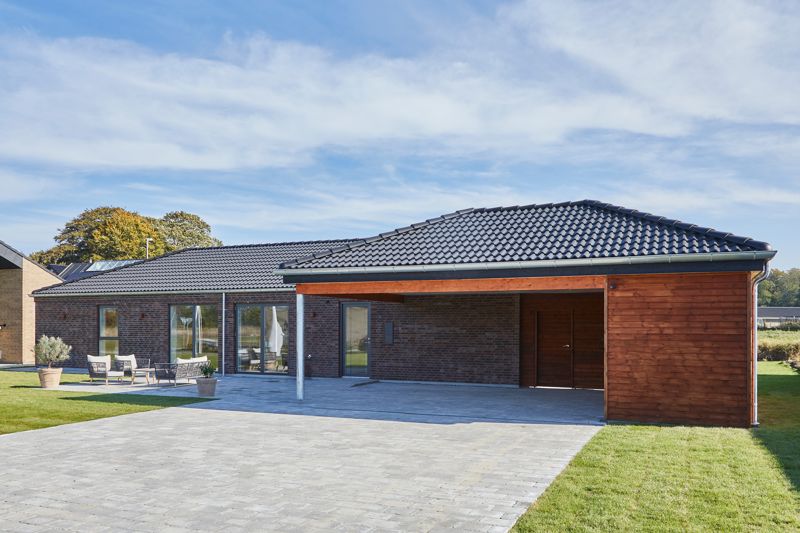 Nybyggeri og husbyggeri med moderne længehus i brunlige mursten, sort tag, træbeklædt carport og overdækket indkørsel fra eurodan-huse