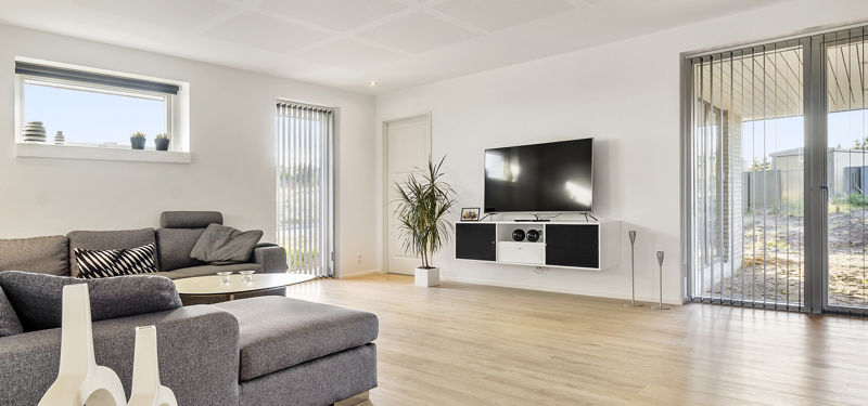 Lys stue fra eurodan-huse med grå hjørnesofa, lyst trægulv, væghængt tv-møbel og store glaspartier, der giver inspiration til moderne husdrømme.