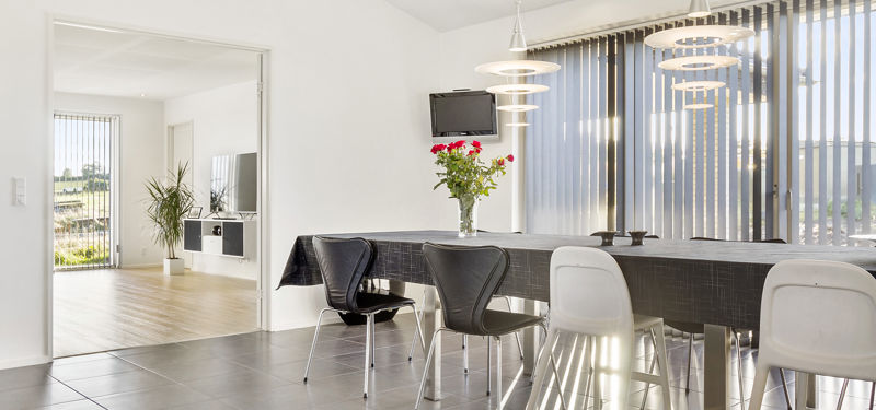Lys spiseplads i et nyt hus fra eurodan-huse med langbord, moderne stole, dekorative pendler og store vinduespartier med lamelgardiner.