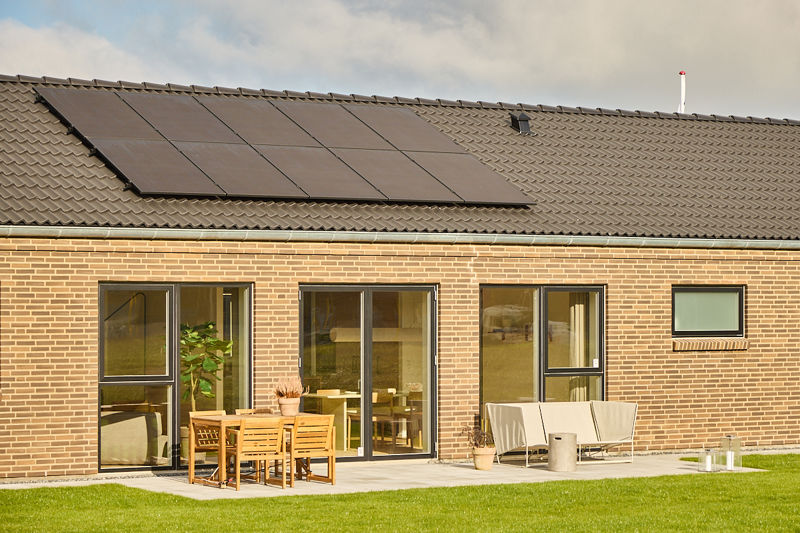 Husbyggeri med lys murstensfacade, sort tegltag, solcellepaneler og terrasse med spiseplads og loungestole foran store glaspartier