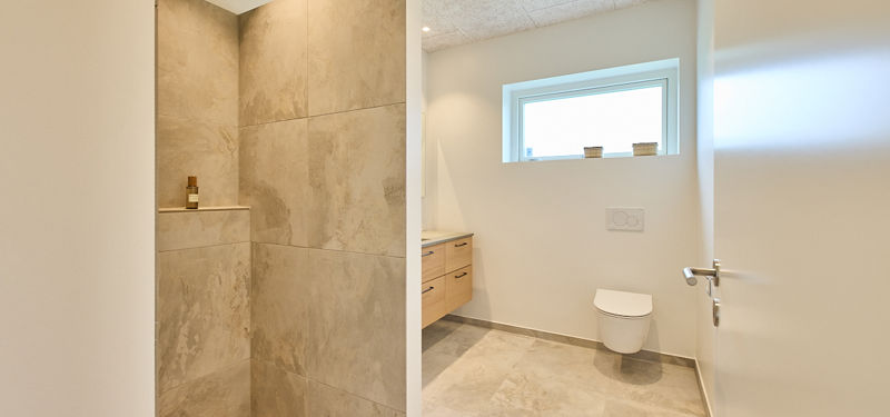Badeværelse i nybygget hus med stor bruseniche, grå fliser, indbygget shampoohylde, væghængt toilet og enkelt nordisk udtryk
