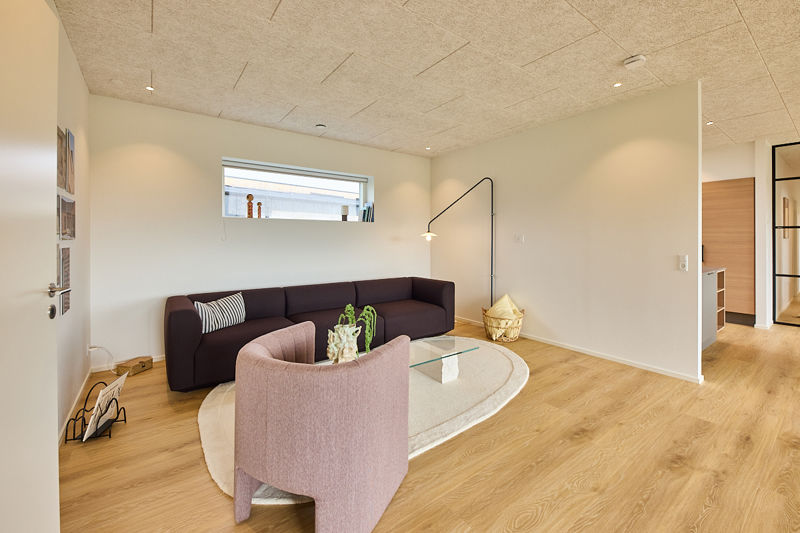Stue fra eurodan-huse med mørk sofa langs væggen, rosa loungestol, ovalt glasbord, højt smalt vindue, sort væglampe og åben forbindelse mod køkken-alrum – konkret inspiration til nybyggeri, husbyggeri og funktionelle husdrømme.Stue fra eurodan-huse med mørk sofa langs væggen, rosa loungestol, ovalt glasbord, højt smalt vindue, sort væglampe og åben forbindelse mod køkken-alrum – konkret inspiration til nybyggeri, husbyggeri og funktionelle husdrømme.