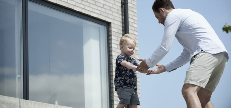 Far leger med sit barn foran et moderne nybygget hus med lyse mursten og store vinduespartier.