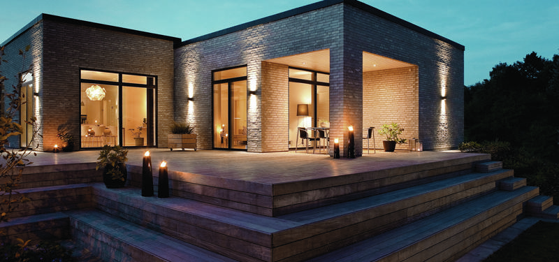 Moderne nybygget hus med fladt tag, lys murstensfacade, store vinduespartier, overdækket terrasse og stemningsfuld udendørsbelysning i skumringen.