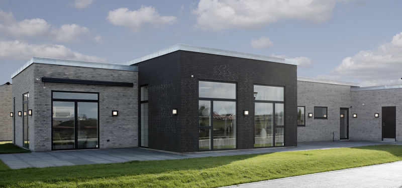 Moderne nybygget hus med fladt tag, mørk og grå murstensfacade, store vinduespartier og grøn græsplæne langs boligen.