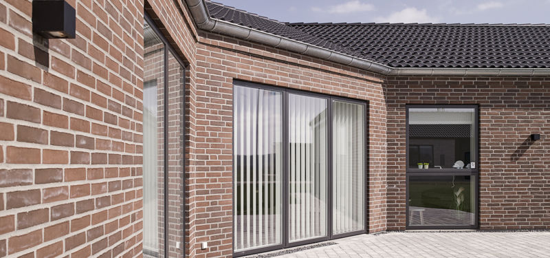 Flisebelagt terrasse ved knækhus på 176 m² med rød murstensfacade, sorte vinduespartier, lodrette lamelgardiner og moderne væglamper – stilren inspiration til nybyg og husdrømme.