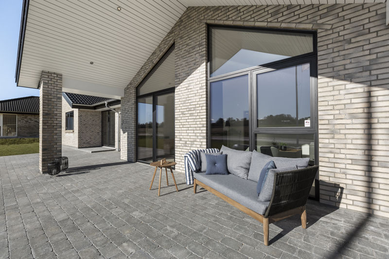 Overdækket terrasse langs lys murstensfacade fra eurodan-huse med store vinduer i mørke rammer, grå fliser, udendørs sofa med puder og lille sidebord under tagudhæng – inspiration til nybyggeri med læfuld terrasse og moderne uderum.
