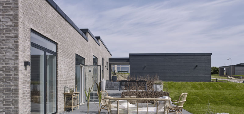 Stor terrasse fra eurodan-huse med mørke fliser, loungeområde i rattan, plantekrukker, store glaspartier, trappetrin i haven og lys murstensfacade med sort stern – inspiration til nybyggeri med rummelig terrasse og afslappede husdrømme.