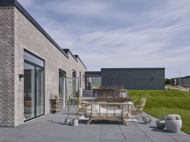 Stor terrasse fra eurodan-huse med mørke fliser, loungeområde i rattan, plantekrukker, store glaspartier, trappetrin i haven og lys murstensfacade med sort stern – inspiration til nybyggeri med rummelig terrasse og afslappede husdrømme.