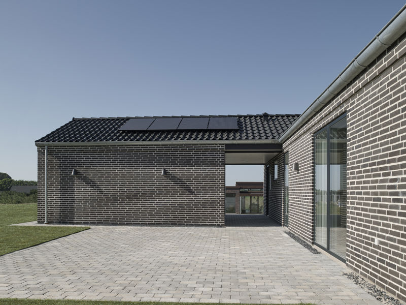 Flisebelagt terrassegård fra eurodan-huse mellem mørke murstensfacader med store vinduespartier, sort tag med solceller og åben passage mod haven – inspiration til nybyggeri med afskærmet uderum og gennemtænkt terrasseindretning.