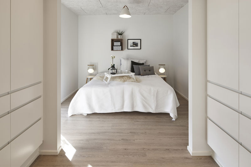 Soveværelse set fra walk-in-område med hvide garderobeskabe på begge sider, brede skuffer med greb, lys dobbeltseng, små sengeborde med lamper og trælook gulv – konkret inspiration til walk-in closet, opbevaring i nybyggeri og gennemtænkte husdrømme fra eurodan-huse.