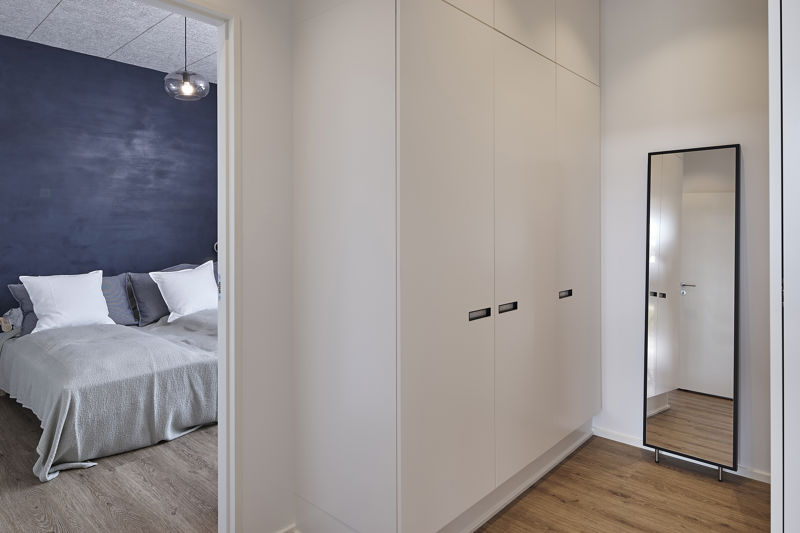 Walk-in closet ved soveværelse med høje hvide skabslåger, sorte greb, fritstående spejl, lyst trægulv og kig ind til seng med mørk blå væg – inspiration til praktisk garderobe, moderne nybyggeri og husbyggeri med god opbevaring fra eurodan-huse.