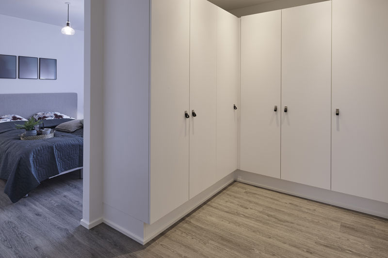 Walk-in closet med hvide skabe fra gulv til loft, hjørneløsning, sorte greb, gråt trælook gulv og åben forbindelse til soveværelse med mørkt sengetæppe – konkret inspiration til garderobeløsning, opbevaring og funktionelt nybyggeri fra eurodan-huse.