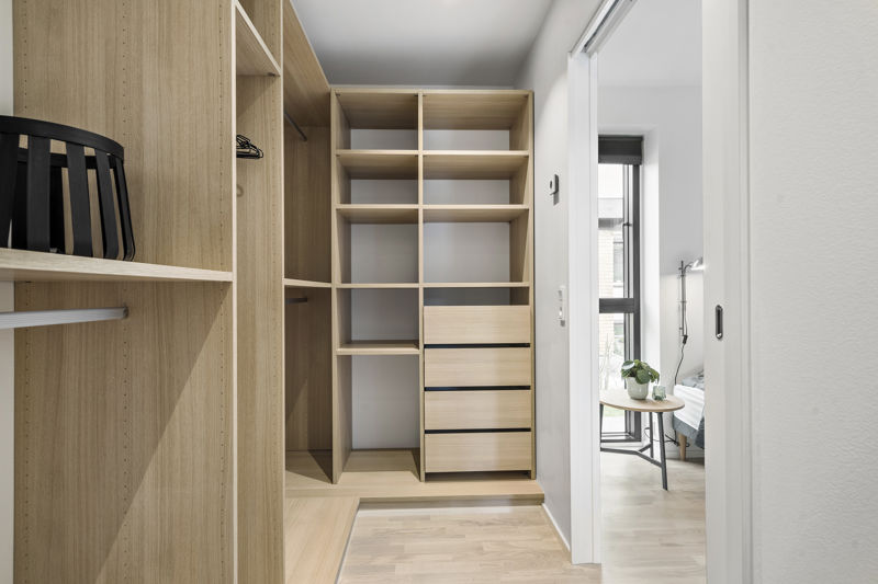 Walk-in closet fra eurodan-huse med skabe i lyst træ, åbne hylder, bøjlestang, skuffer, sort taske på hylde og kig gennem skydedør til soveværelse med lille bord og plante – inspiration til varm og praktisk garderobe i nybygget hus.