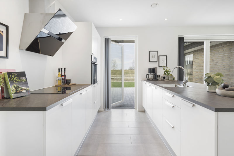 Køkken fra eurodan-huse med lange hvide køkkenelementer, mørk bordplade, markant emhætte i metal, integreret ovn, stort vindue over vasken og åben dør ud mod terrasse og have – inspiration til vinduer, døre og lyse køkkenløsninger i nybyggeri.