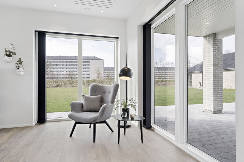 Læsehjørne fra eurodan-huse med stor gulv-til-loft vinduesløsning, hjørnevindue mod overdækket terrasse, grå lænestol, rundt sidebord og sort gulvlampe – inspiration til vinduer, lysindfald og hyggelige rum i nybyggeri.