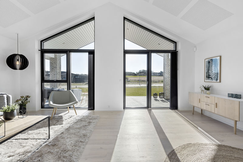 Lyst opholdsrum fra eurodan-huse med to høje gavlvinduer, sorte rammer, terrassedøre, hvide vægge, lys lænestol, lav skænk og markant loftshøjde – inspiration til store vinduespartier, dagslys og nybyggeri med åbne opholdsrum.