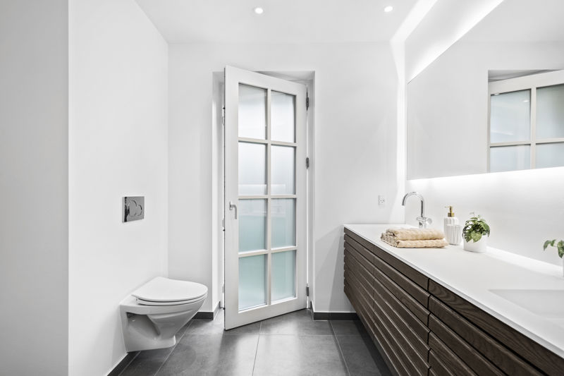 Badeværelse fra eurodan-huse med hvid dør med matterede glasfelter, væghængt toilet, mørkt badmøbel med mange skuffer, hvid bordplade, stort spejl og indbygget belysning – inspiration til indvendige døre, lysindfald og funktionelt nybyggeri.