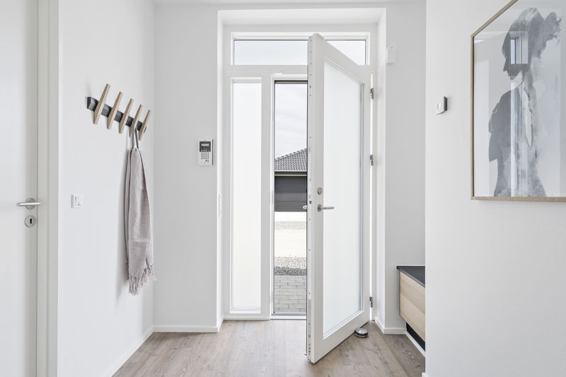 Lys entré fra eurodan-huse med åben hoveddør, høje sidevinduer, trælook gulv, vægmonteret knagerække, bænk med mørk hynde og stor vægplakat – inspiration til funktionel indgang i nybyggeri og husdrømme.