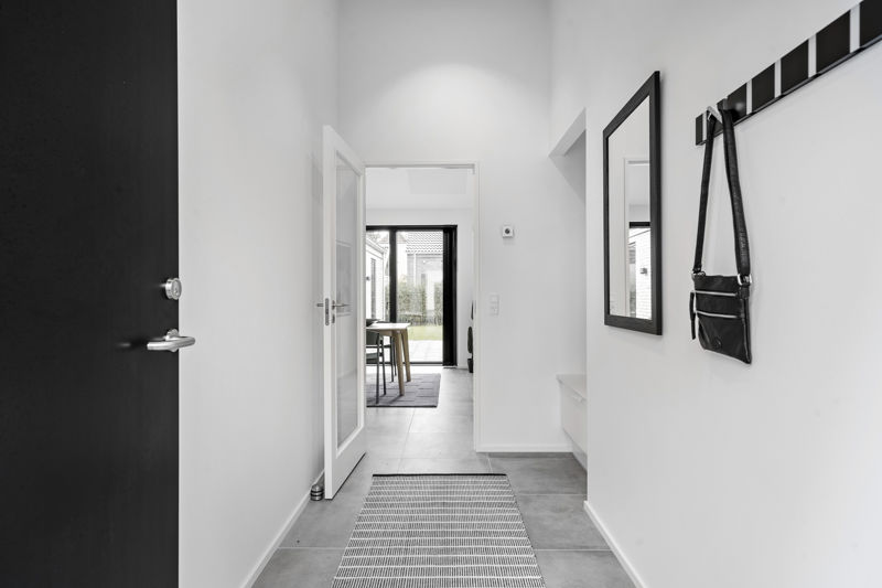 Smal entré fra eurodan-huse med sort hoveddør, grå fliser, lang gulvløber, sort spejl, knagerække med taske og åben dør mod køkken-alrum – inspiration til kompakt entré, nybyggeri og moderne husdrømme.